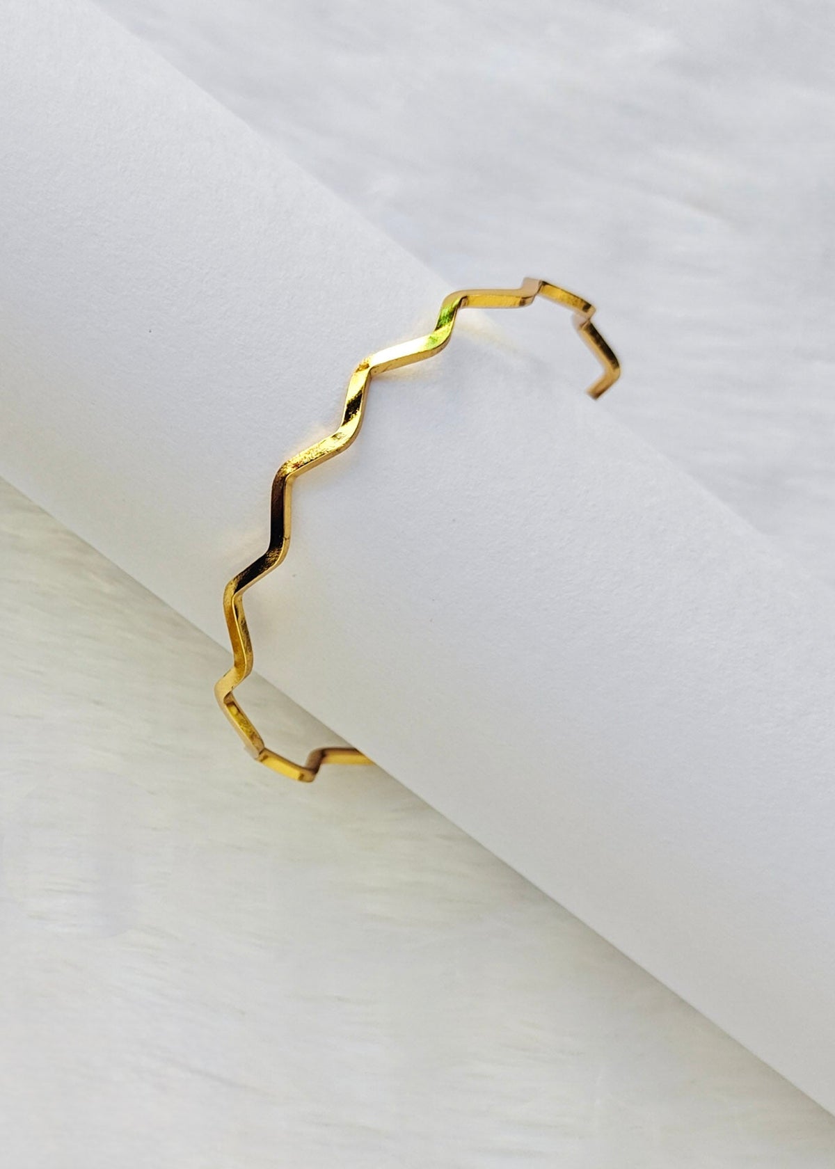 A thin, open cuff bracelet with a wavy, zigzag gold-plated edge, displayed on a white textured surface.