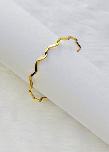A thin, open cuff bracelet with a wavy, zigzag gold-plated edge, displayed on a white textured surface.