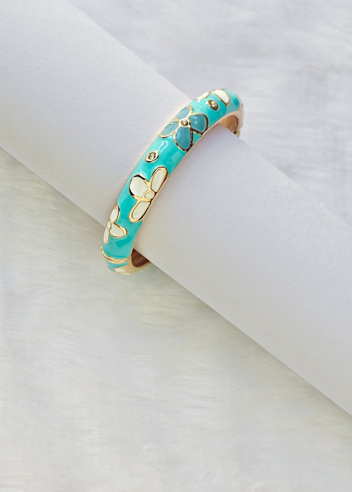 A turquoise and gold butterfly cuff bracelet displayed on a white cylindrical stand, with a gold beaded necklace partially visible in the lower-left corner on a white textured background.