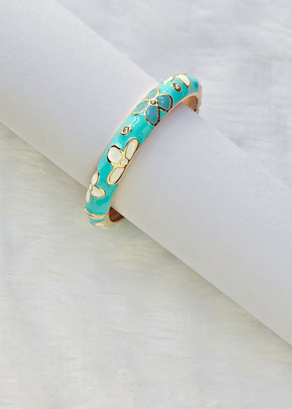 A turquoise and gold butterfly cuff bracelet displayed on a white cylindrical stand, with a gold beaded necklace partially visible in the lower-left corner on a white textured background.