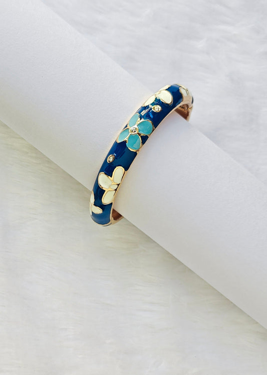 A gold-tone bangle bracelet with a navy blue floral enamel design, displayed on a white cylindrical stand against a white textured background.
