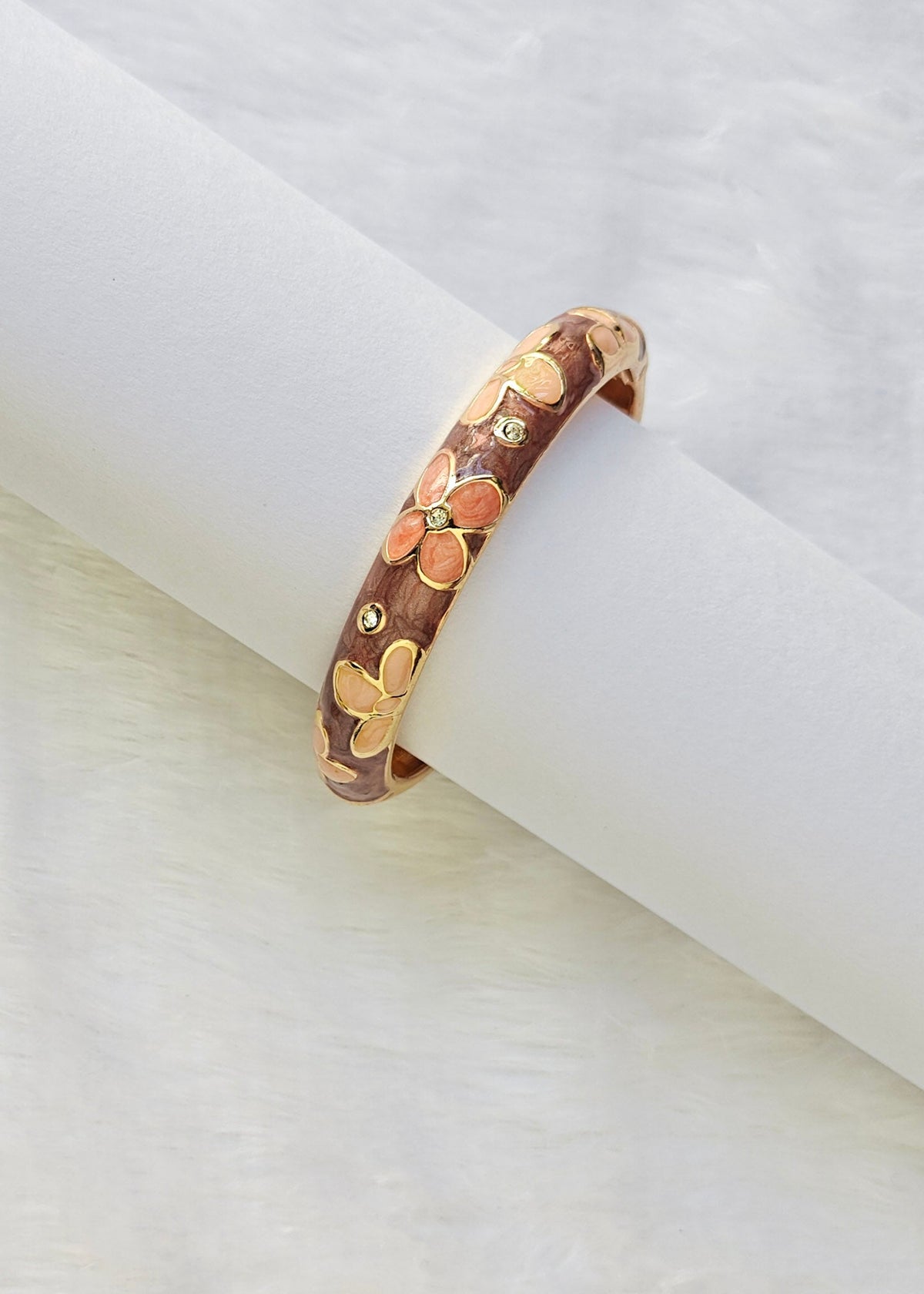 A gold-plated bangle bracelet with a brown floral enamel pattern, displayed on a white cylindrical stand against a white textured background.