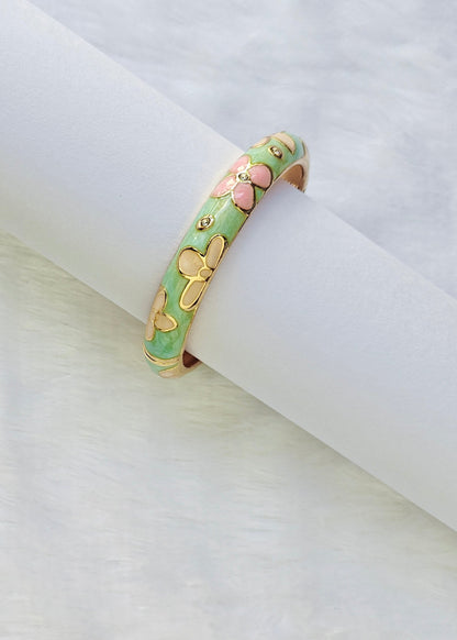 A gold-plated bangle bracelet with a mint green enamel background and pink and gold butterfly motifs, displayed on a white cylindrical stand.