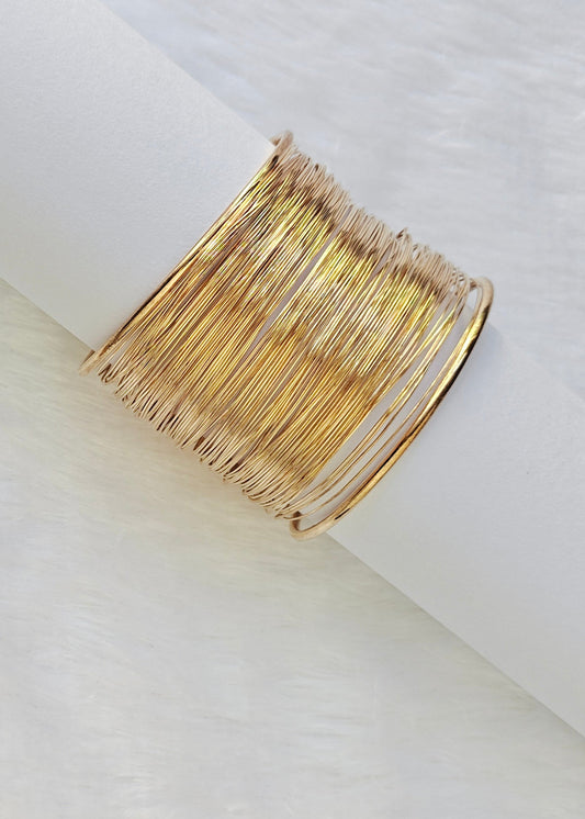 A wide gold-toned wire cuff bracelet displayed on a white cylindrical stand against a soft white background.