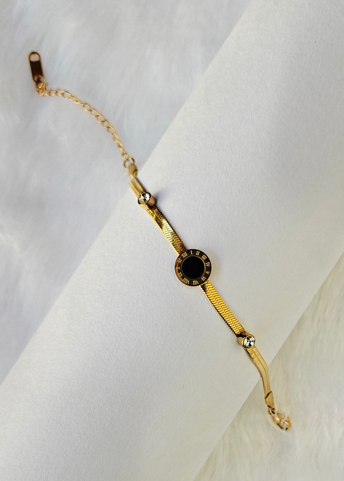 A gold chain bracelet with a central round black stone and small clear accent stones, displayed on a white textured background.