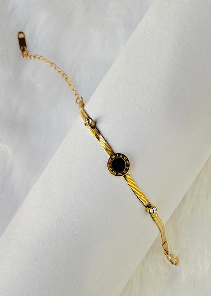 A gold chain bracelet with a central round black stone and small clear accent stones, displayed on a white textured background.