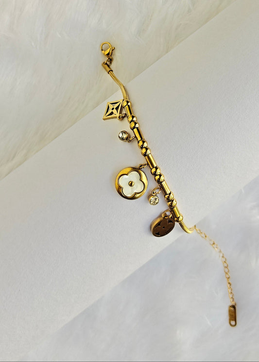A gold-tone charm bracelet featuring a central flower charm, a small clover motif, and a textured bar, displayed on a white cylindrical stand against a white faux fur background.