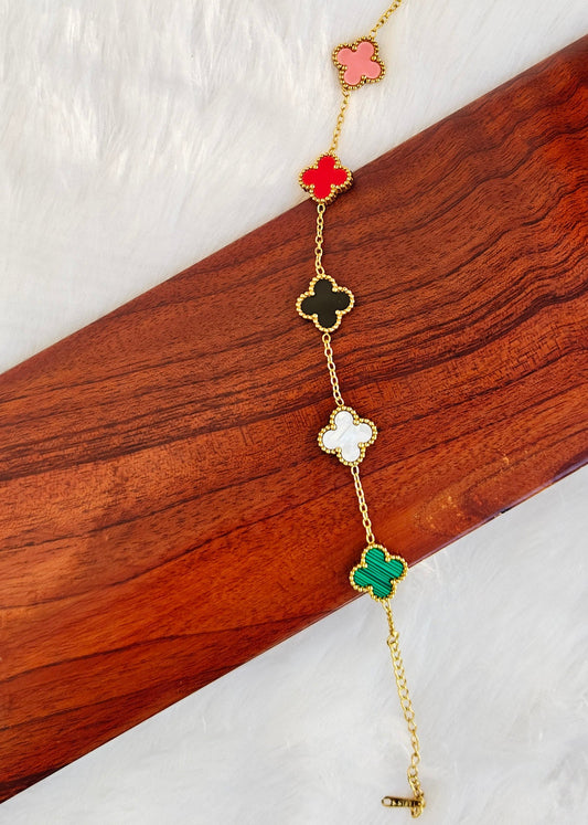 A gold-tone chain bracelet with five small clover-shaped links in pink, red, black, white, and green, displayed on a diagonal wooden block over a white textured background.