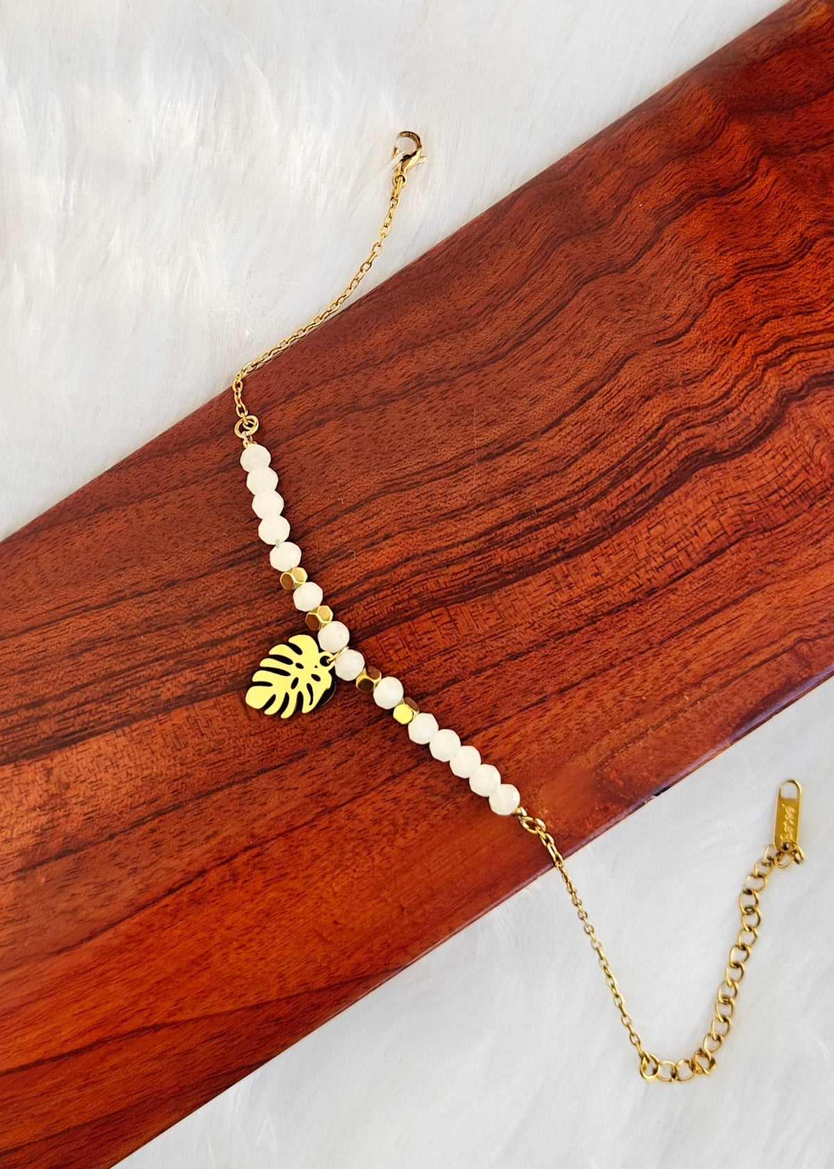 A gold chain bracelet featuring white pearl-like beads and a gold monstera leaf charm, displayed on a dark wood block over a white faux fur background.