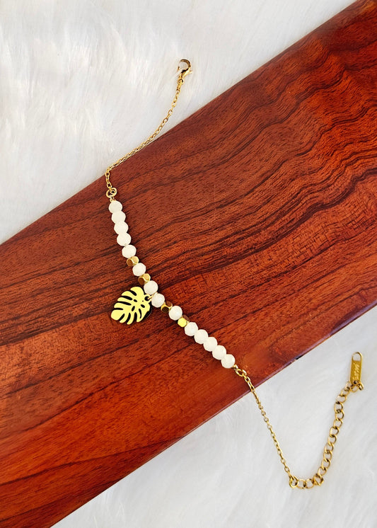 A gold chain bracelet featuring white pearl-like beads and a gold monstera leaf charm, displayed on a dark wood block over a white faux fur background.