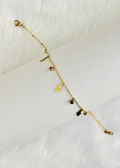 A delicate gold chain bracelet with small dangling charms, including a heart and tiny dark and red stones, displayed on a white cylindrical stand against a white faux fur background.