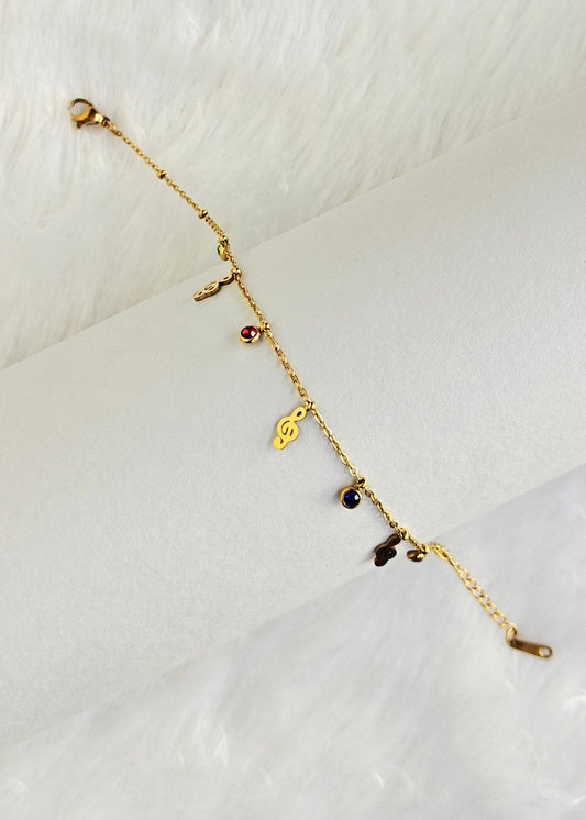A delicate gold chain bracelet with small dangling charms, including a heart and tiny dark and red stones, displayed on a white cylindrical stand against a white faux fur background.