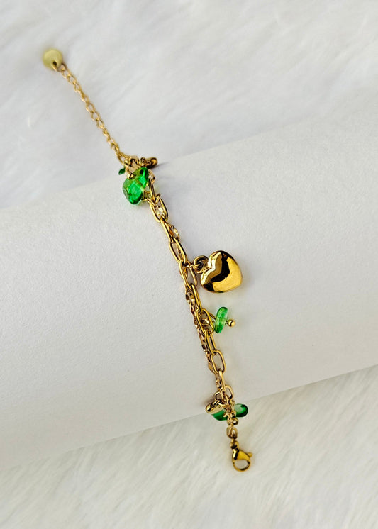 A delicate gold chain bracelet with small green crystal charms and a polished gold heart pendant, displayed on a white cylindrical stand against a white faux fur background.