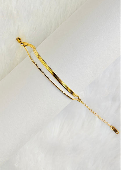 A delicate gold chain bracelet with a polished curved bar and a fine chain link overlay, displayed on a white textured surface.
