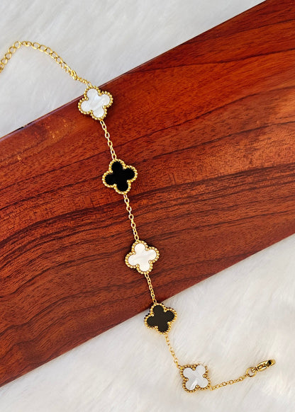 A gold-tone chain bracelet with five clover motifs, alternating between black and white enameled centers, displayed diagonally on a split background of dark wood and white faux fur.