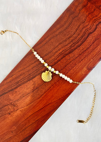 A gold chain bracelet with white pearl-like beads, a scallop shell charm, and a lobster clasp, displayed on a diagonal wooden board over a white textured background.