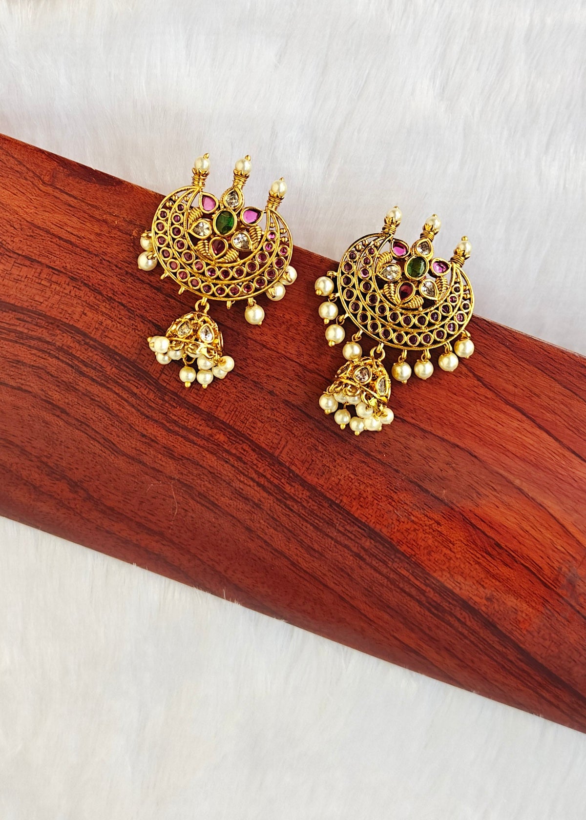 A pair of ornate gold-toned Indian jhumka earrings with colorful gemstone accents and dangling pearl beads, displayed on a polished wooden block.