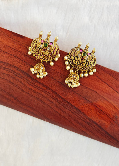 A pair of ornate gold-toned Indian jhumka earrings with colorful gemstone accents and dangling pearl beads, displayed on a polished wooden block.