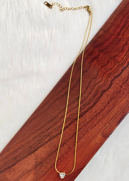 A delicate gold chain necklace with a small heart-shaped clear stone pendant, displayed on a diagonal dark reddish-brown wooden board over a white textured background.