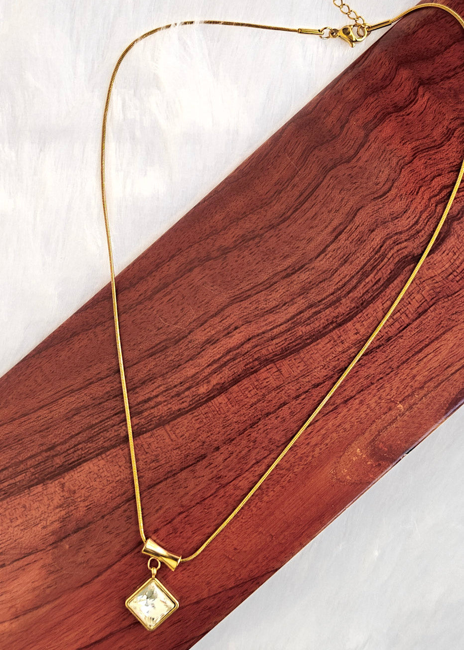 A delicate gold chain necklace with a small square pendant, displayed on a dark wood block over a white textured background.