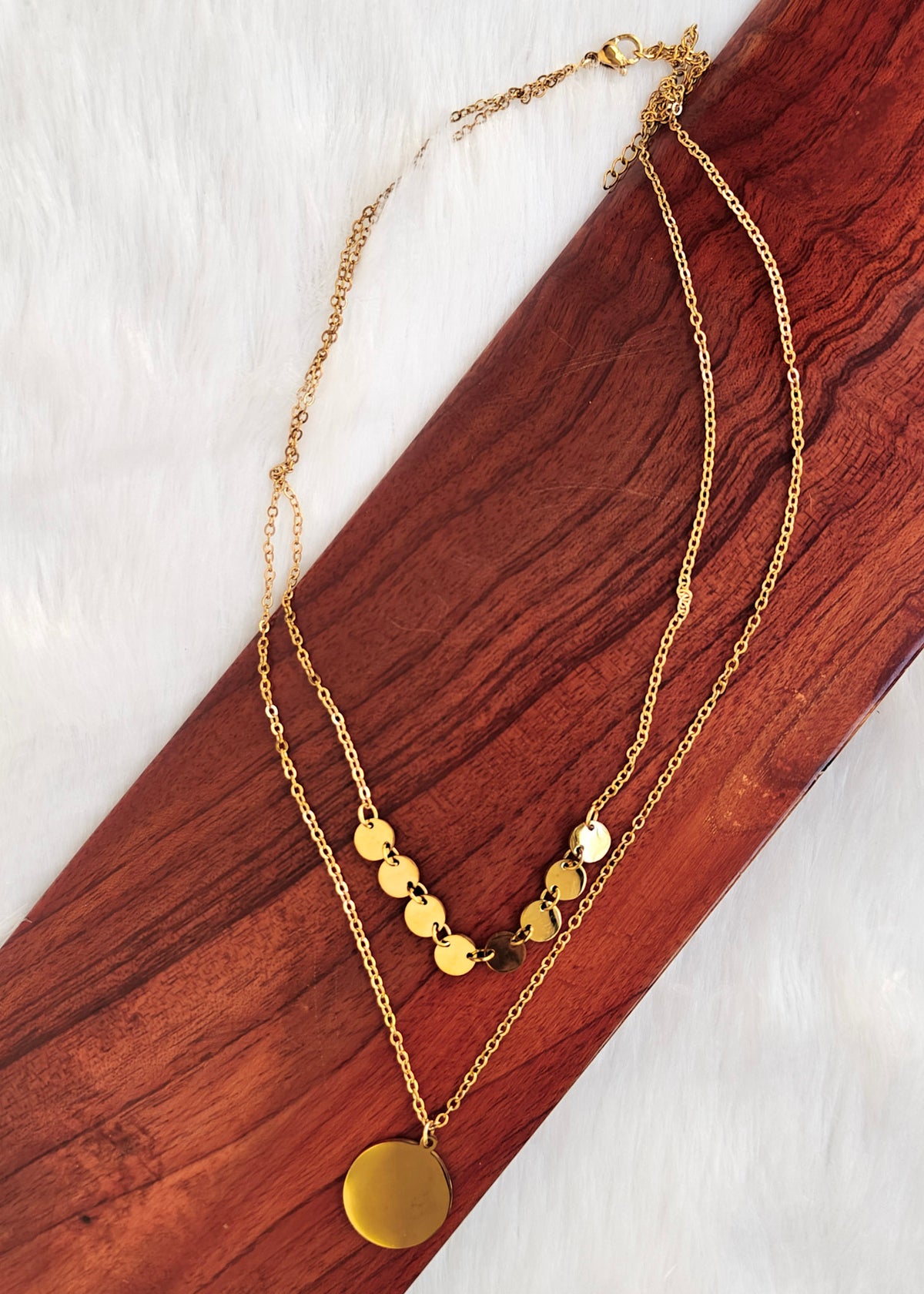 A gold-tone anti tarnish double-layer necklace with small round disc charms and a larger circular pendant, displayed on a diagonal wooden board over a white faux-fur background.