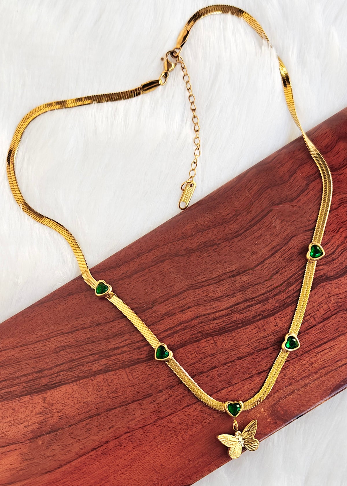 Anti tarnish Gold snake chain necklace with green heart-shaped accents and a butterfly pendant, displayed on a wooden block over a white textured background.