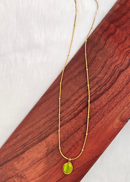 A gold chain necklace with a small oval lime green pendant, displayed on a diagonal wooden block over a white textured background.