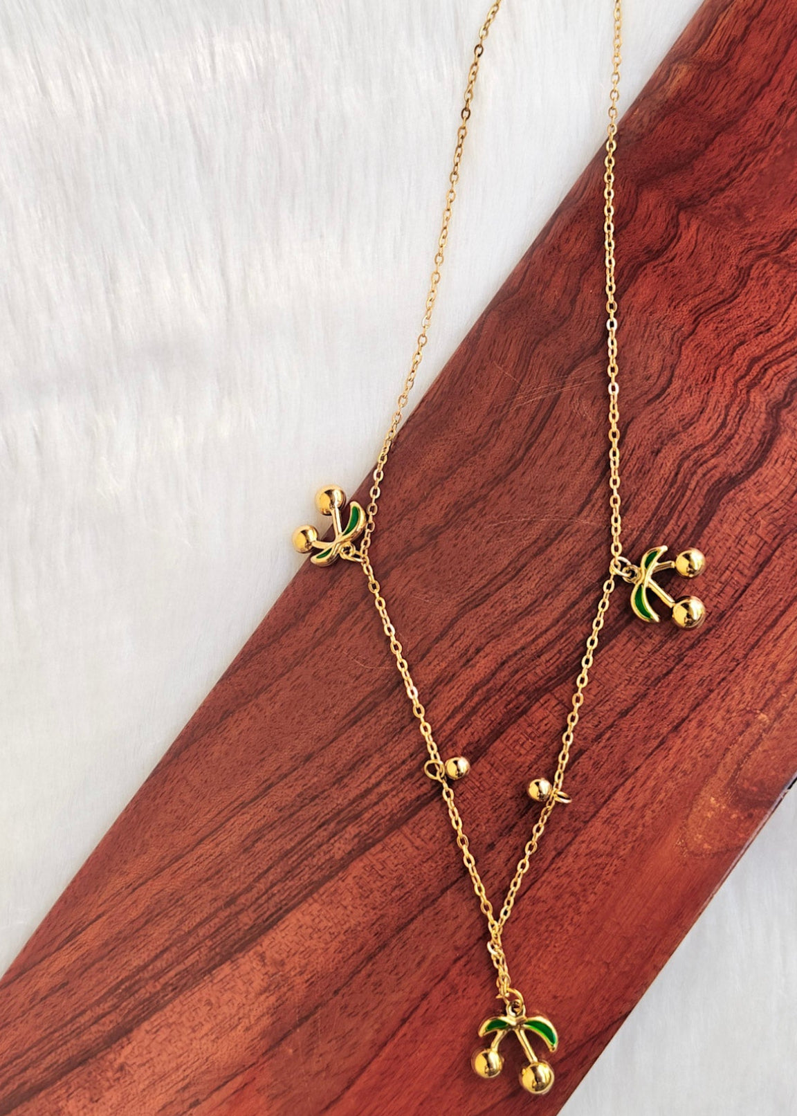 A gold chain necklace with three small gold leaf pendants and a larger gold leaf pendant, displayed on a diagonal wooden block over a white textured background.