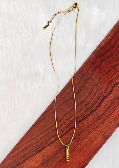 A delicate anti tarnish gold chain necklace with a small vertical bead drop pendant, displayed on a diagonal dark wood block over a white faux fur background.