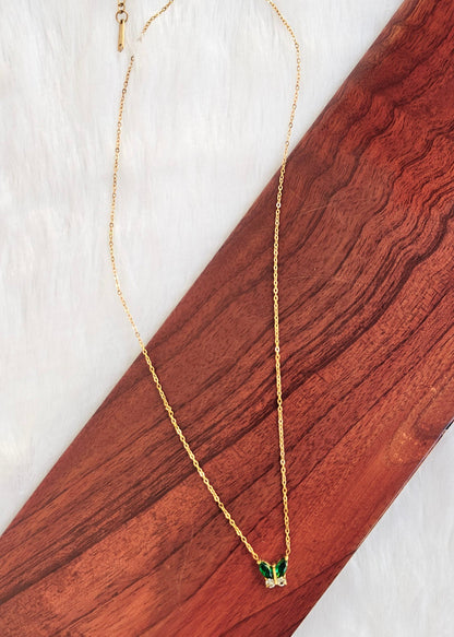 A delicate gold chain necklace with a small butterfly pendant featuring green wings and clear stones, displayed on a diagonal wooden block over a white textured background.