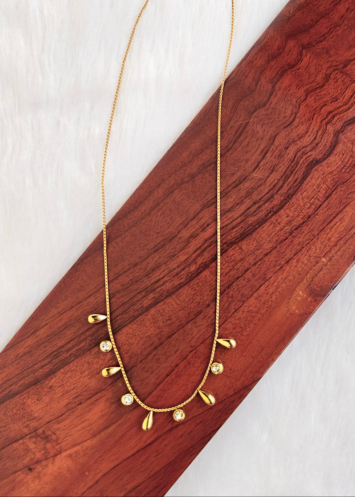 A delicate gold chain necklace with small teardrop-shaped gold charms and clear rhinestone accents, displayed on a diagonal wooden block over a white textured background.