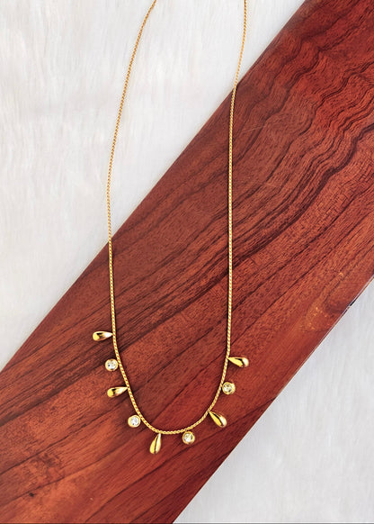 A delicate gold chain necklace with small teardrop-shaped gold charms and clear rhinestone accents, displayed on a diagonal wooden block over a white textured background.