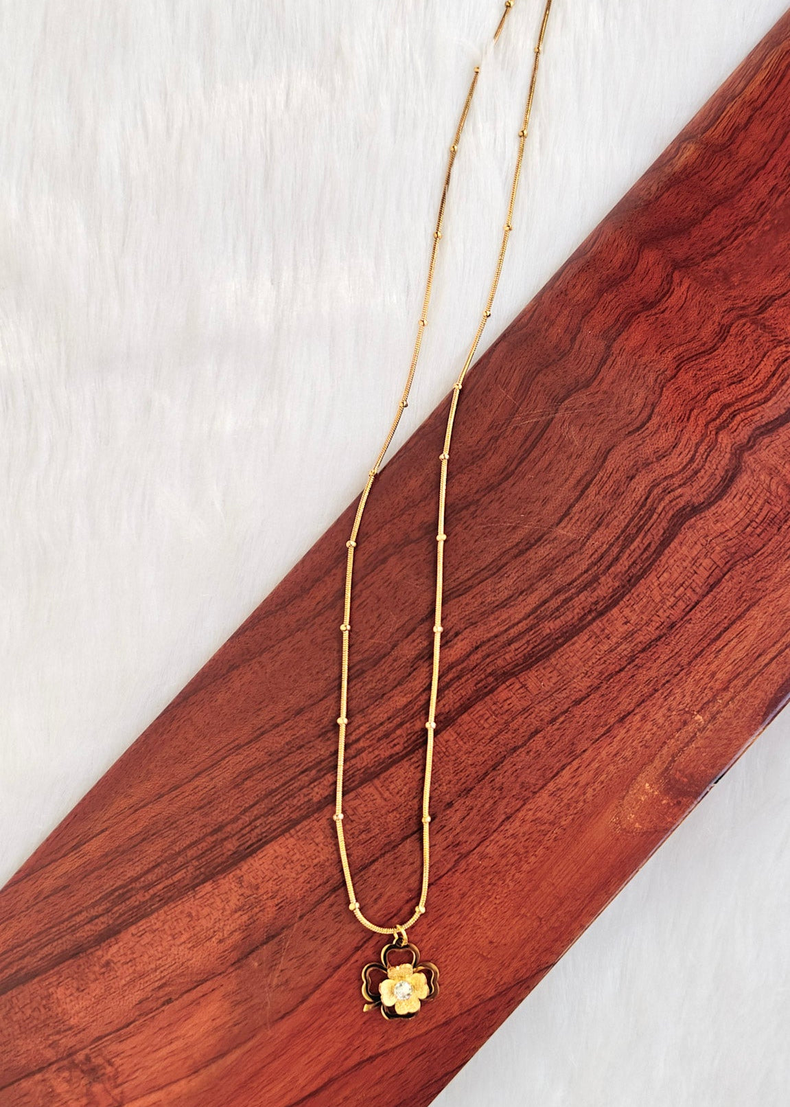 A gold-toned double-strand necklace with a small flower-shaped pendant, laid diagonally on a dark reddish-brown wooden block over a white textured background.