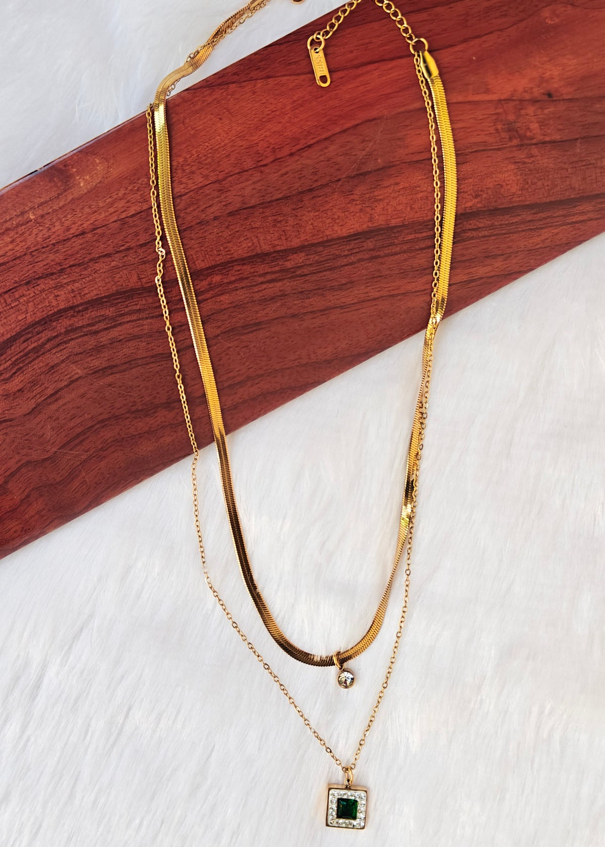 A double-layered anti tarnish gold necklace with a flat chain and a delicate chain featuring a square pendant with a green center stone,
