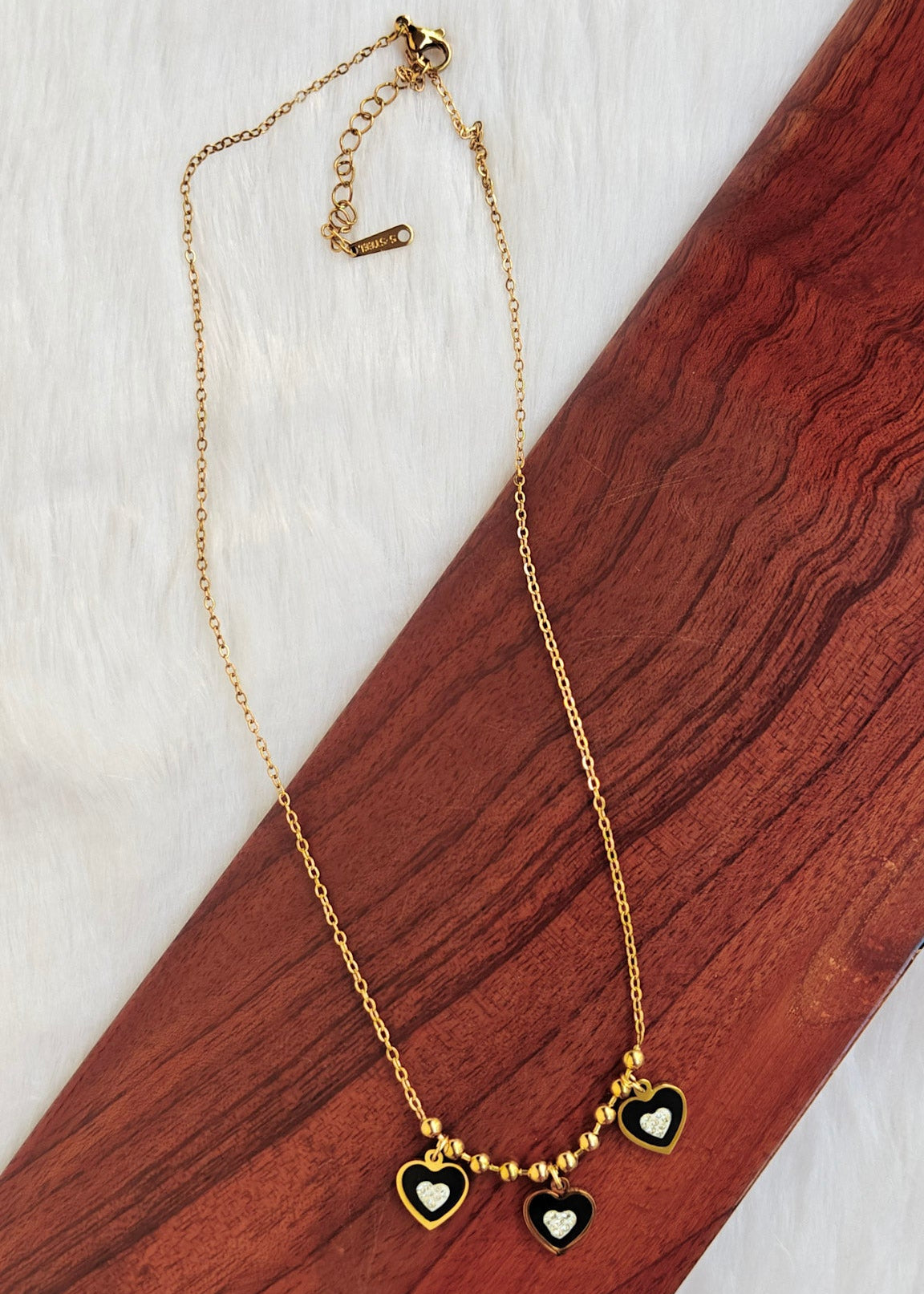 A gold chain necklace with three small heart charms, two of which are black with white centers, displayed on a diagonal wooden block over a white textured background.