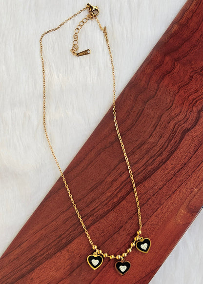 A gold chain necklace with three small heart charms, two of which are black with white centers, displayed on a diagonal wooden block over a white textured background.