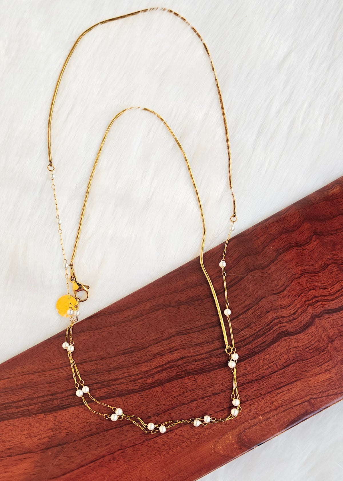 A delicate gold chain necklace with small white pearl-like beads and a small yellow circular charm, displayed on a dark wood block against a white textured background.