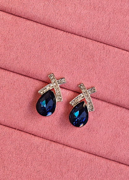 A pair of silver-toned teardrop stud earrings with a small cross-shaped top set with clear crystals and a deep blue faceted teardrop crystal below, displayed on a pink textured fabric background.