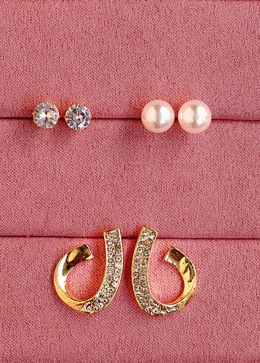 Three pairs of stud earrings displayed on a pink textured jewelry box: two pairs with clear round stones and one pair with white pearl-like beads, plus a pair of gold hoop earrings with clear stones, arranged on a pink velvet-lined box.