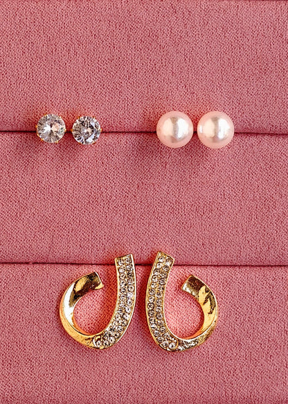 Three pairs of stud earrings displayed on a pink textured jewelry box: two pairs with clear round stones and one pair with white pearl-like beads, plus a pair of gold hoop earrings with clear stones, arranged on a pink velvet-lined box.