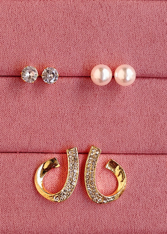 Three pairs of stud earrings displayed on a pink textured jewelry box: two pairs with clear round stones and one pair with white pearl-like beads, plus a pair of gold hoop earrings with clear stones, arranged on a pink velvet-lined box.