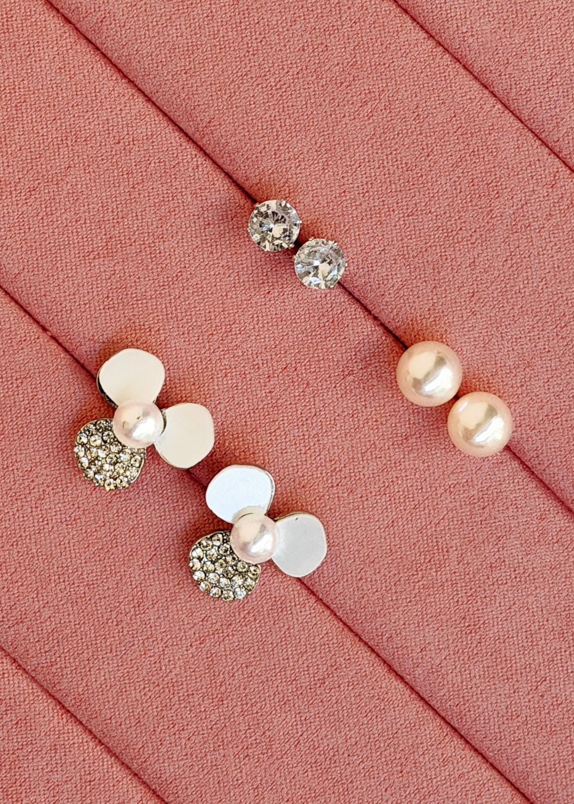 Three pairs of stud earrings displayed on a pink fabric jewelry organizer. The earrings feature white faux pearls, clear rhinestone-covered round studs, and a cluster design combining white discs, clear rhinestones, and a pearl.