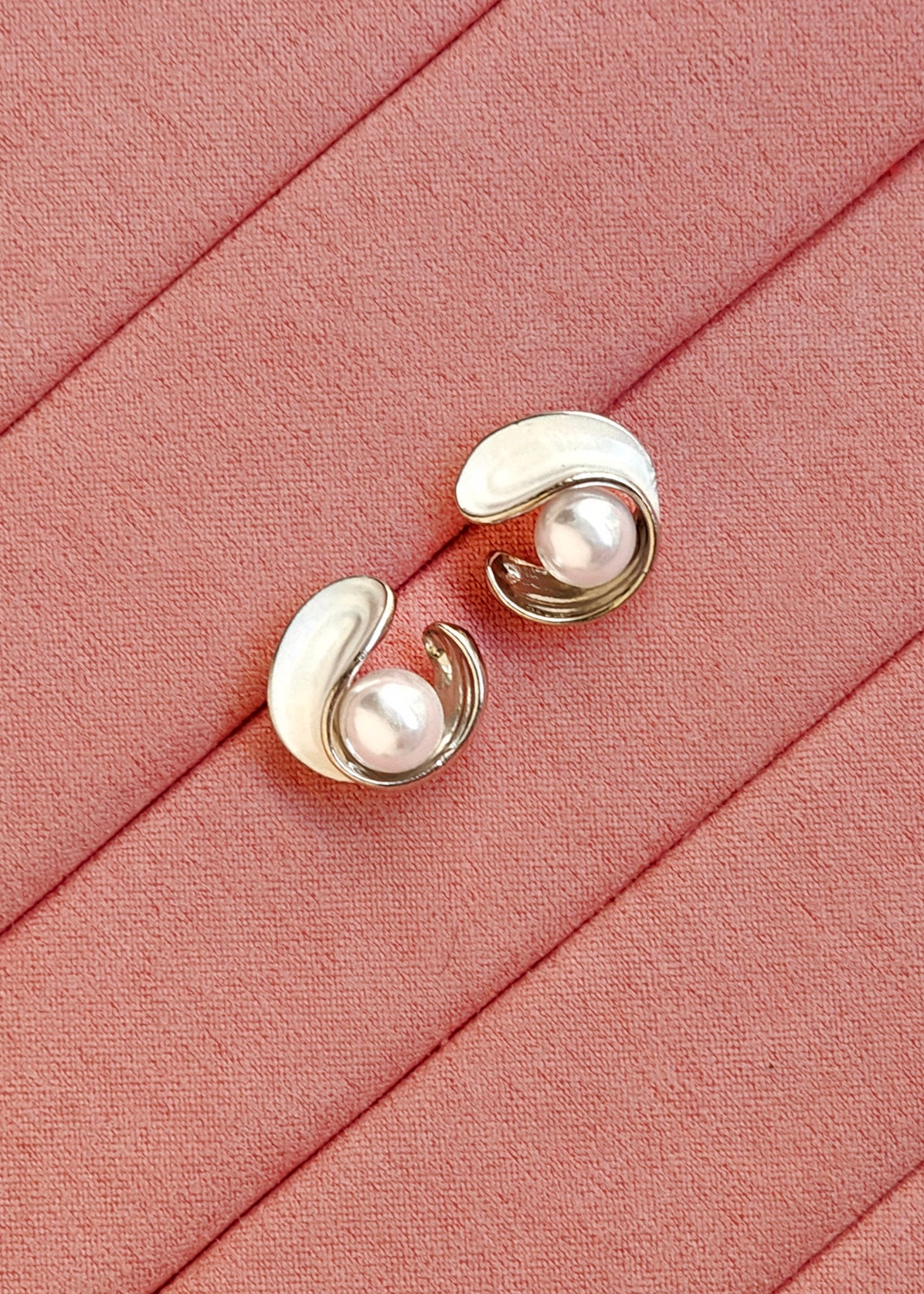 A pair of gold and white enamel curved stud earrings with pearl-like centers, displayed on a textured pink fabric background.