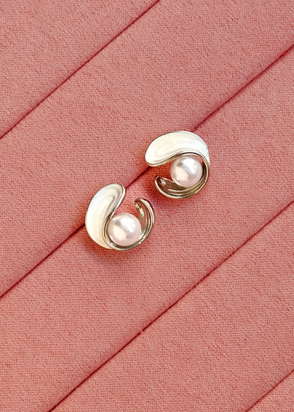 A pair of gold and white enamel curved stud earrings with pearl-like centers, displayed on a textured pink fabric background.