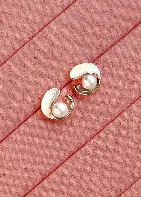 A pair of gold and white enamel curved stud earrings with pearl-like centers, displayed on a textured pink fabric background.