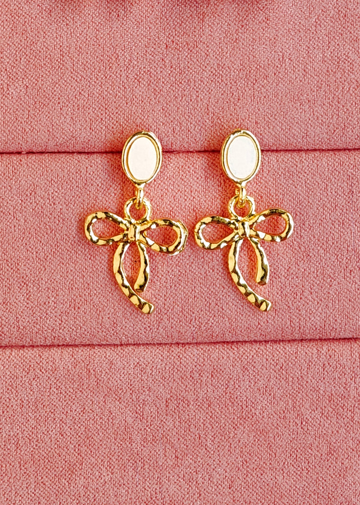 A pair of gold bow-shaped drop earrings with oval white inlays, displayed on a textured pink fabric background.