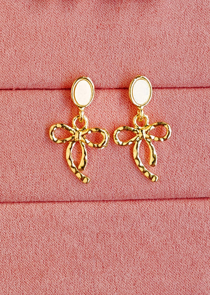 A pair of gold bow-shaped drop earrings with oval white inlays, displayed on a textured pink fabric background.