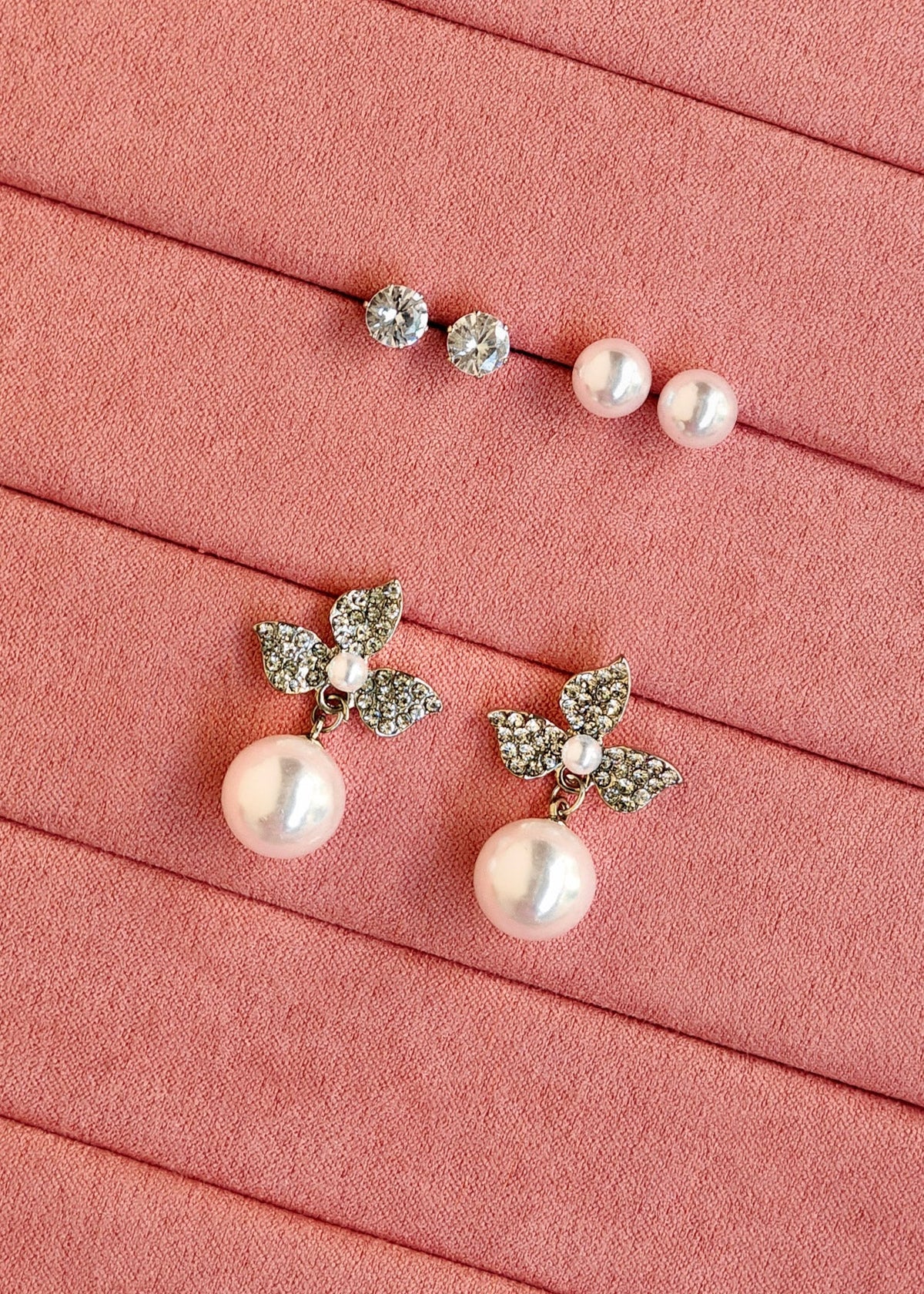 A pair of pearl drop earrings with silver leaf-shaped settings and a matching pair of stud earrings with small clear stones and pearls, arranged on a pink textured background.