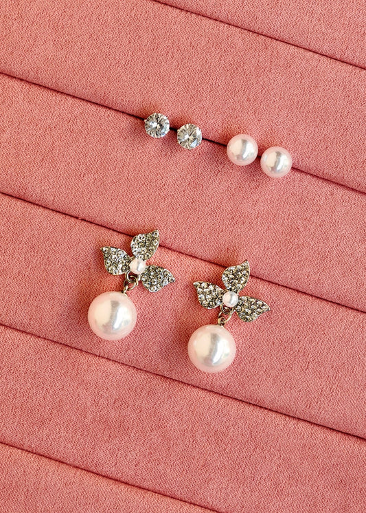 A pair of pearl drop earrings with silver leaf-shaped settings and a matching pair of stud earrings with small clear stones and pearls, arranged on a pink textured background.