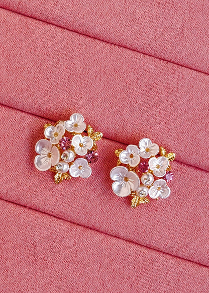 A pair of floral stud earrings with white mother-of-pearl flowers, pink accents, and gold details, displayed on a textured pink fabric background.